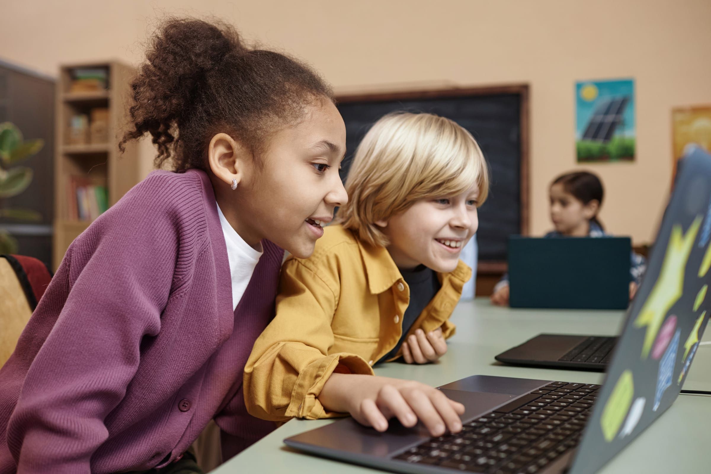 Two children smiling at a laptop in a classroom